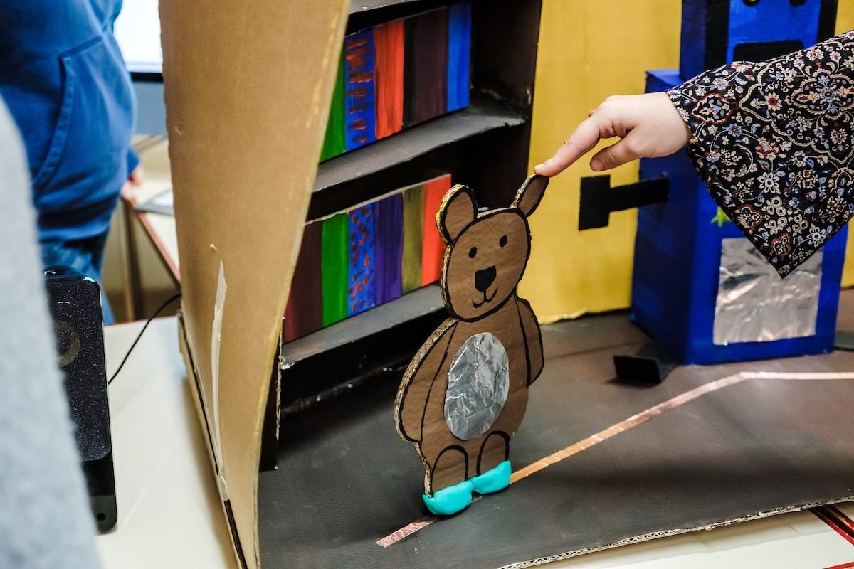 A hand touches the ear tip of a brown cardboard bear figure with a silver belly and blue feet against a colorful background.