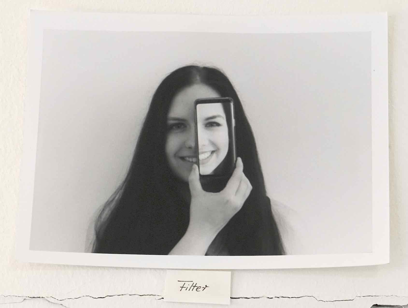 Photograph of a black and white photo on a wall: a person with long dark hair holds a smartphone in front of their face