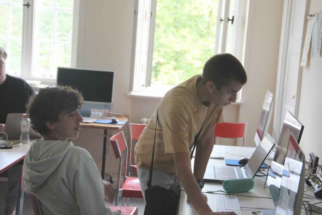 Ein Junge sitzt vor einem Rechner, ein anderer Junge steht daneben. Beide schneiden am Rechner einen Dokumentarfilm zum Seminar