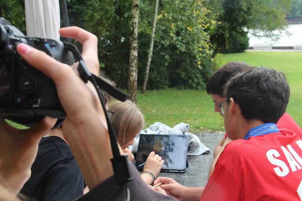 Drei Jugendliche arbeiten an einem iPad an ihrem meme-Projekt an einem Tisch auf der Terrasse. Eine vierte Jugendliche filmt sie dabei für den Dokumentarfilm