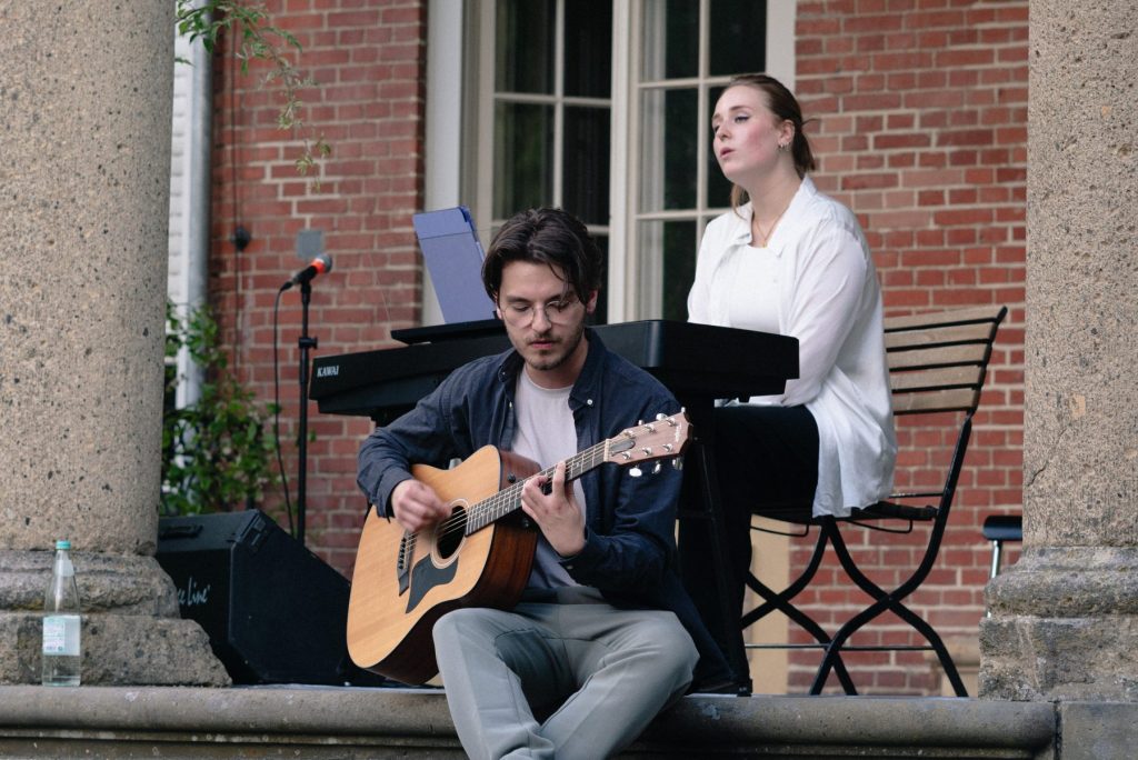 Eine junge Frau sitzt auf der Veranda an einem Keyboard und singt. Vor ihr sitzt ein junger Mann und spielt eine akustische Gitarre.