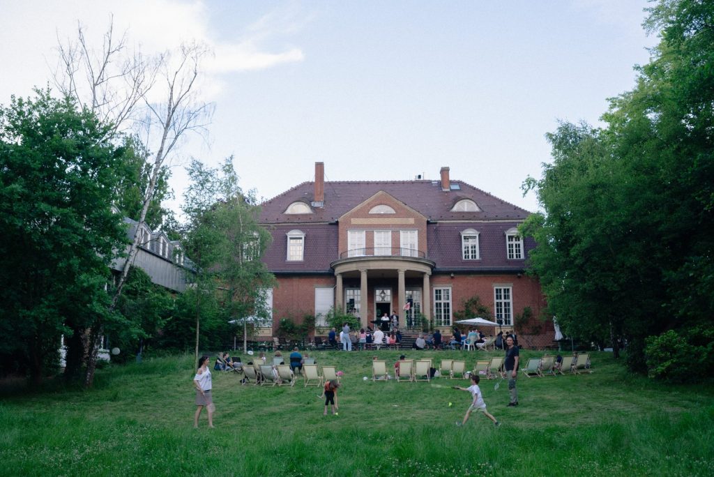 Eine Großaufnahme des Landhauses. Vor dem Haus sieht man einige Menschen tanzen und Federball spielen.