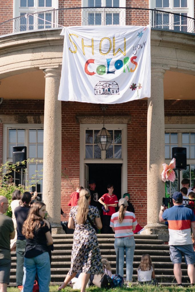 Auf der Veranda eines Hauses stehen drei Kinder. Über ihnen hängt ein Banner mit der Aufschrift "Show your colors". Ihnen schauen mehrere Menschen vor dem Haus zu.