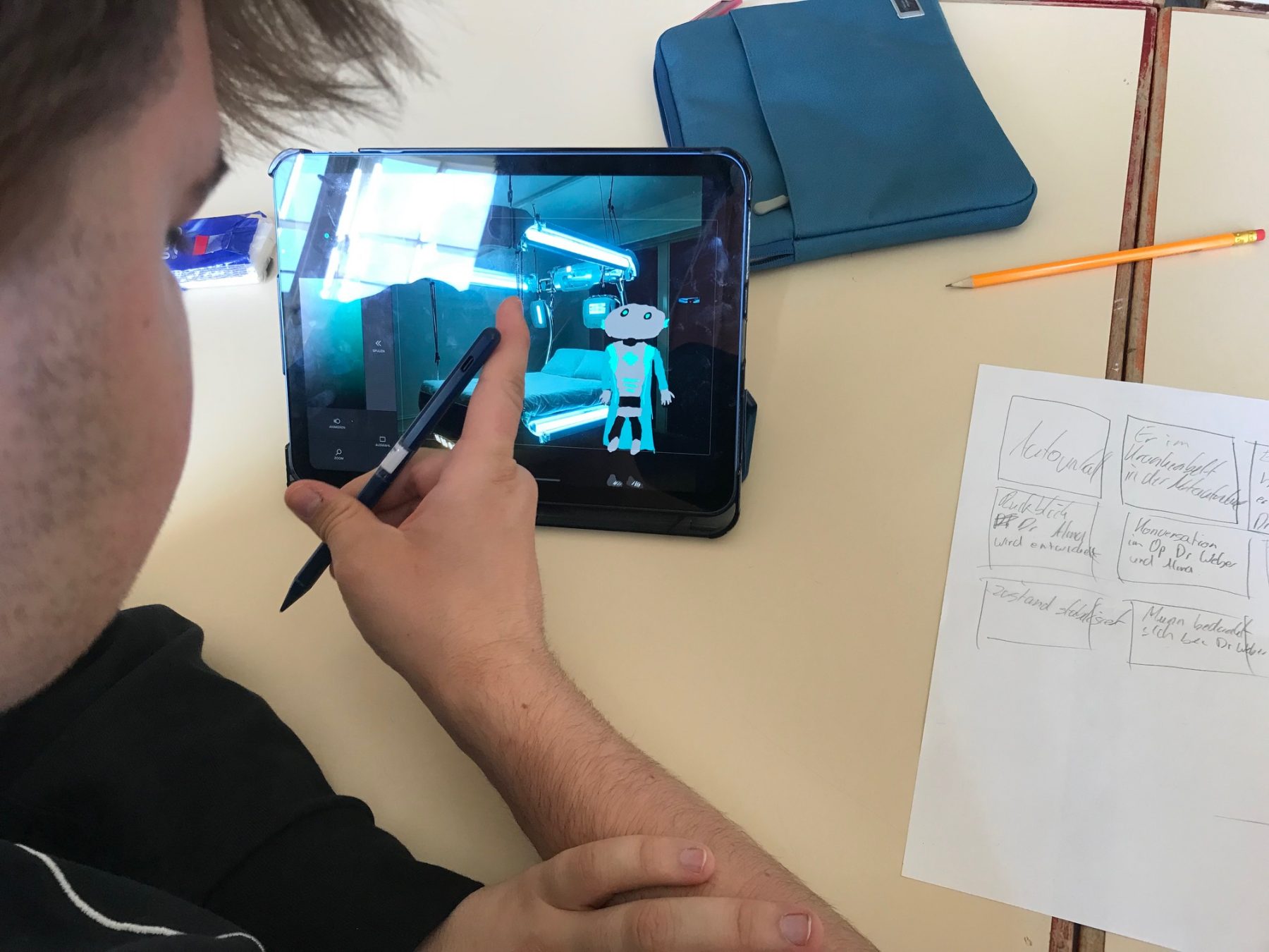 Person zeichnet mit einem Stylus auf einem Tablet einen blauen Roboter, daneben liegen ein Bleistift, ein blauer Mäppchen und ein Blatt Papier mit handschriftlichen Notizen