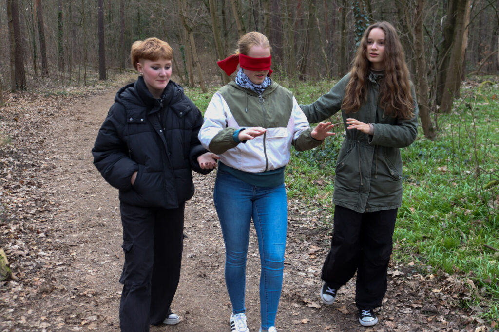 Three teenagers on a forest path, one of them is blindfolded in a trust exercise and is led by the others