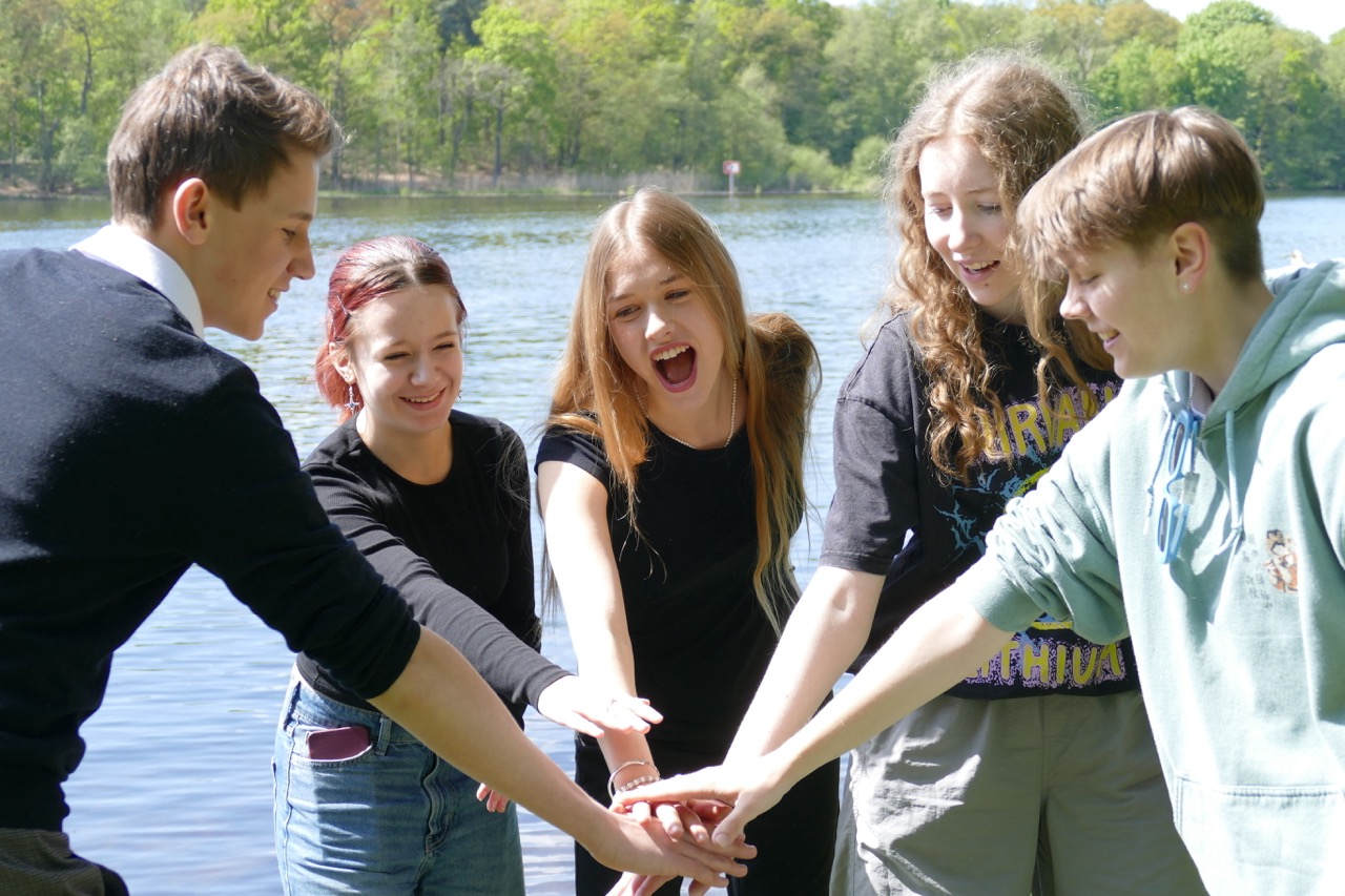 Five teenagers stand in a circle on the lakeshore and place their hands in the middle as a sign of team spirit