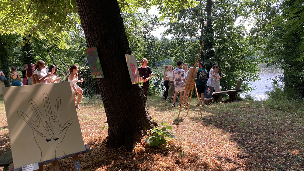 Menschen betrachten selbst gemalte Kunstwerke auf Staffeleien im Freien unter Bäumen neben einem See 