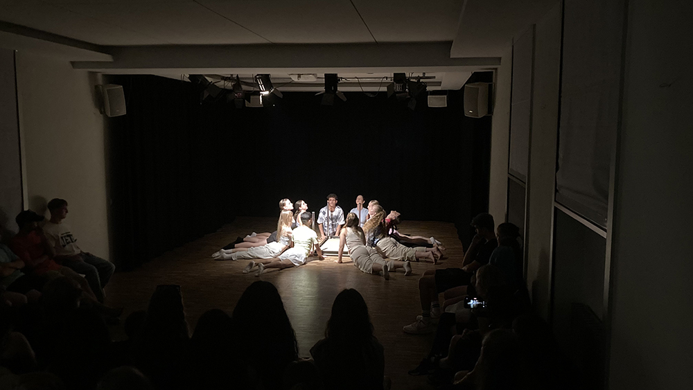 Szenische Theateraufführung mit sieben jungen Schauspieler*innen in weißen Kostümen auf dunkler Bühne, Publikum im Halbdunkel