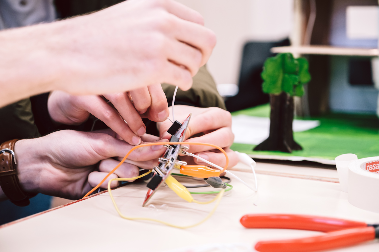Hands connect electronic components with cables on a table, in the background a model tree