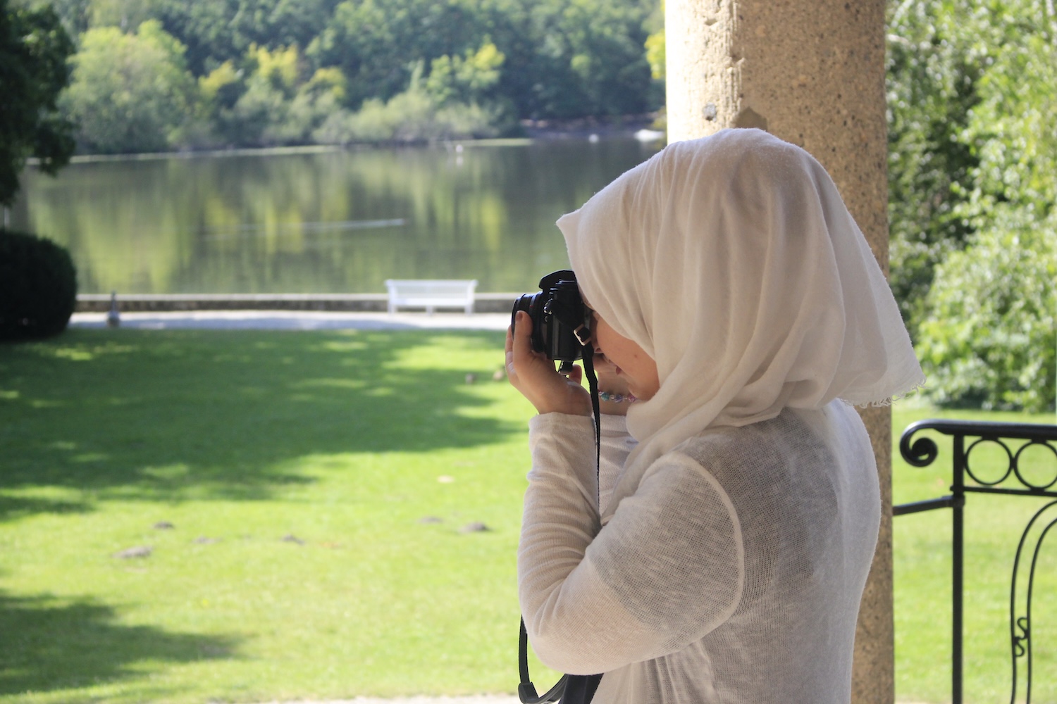 Mädchen mit hellem Kopftuch fotografiert mit Kamera an einem See mit grüner Wiese und Bäumen