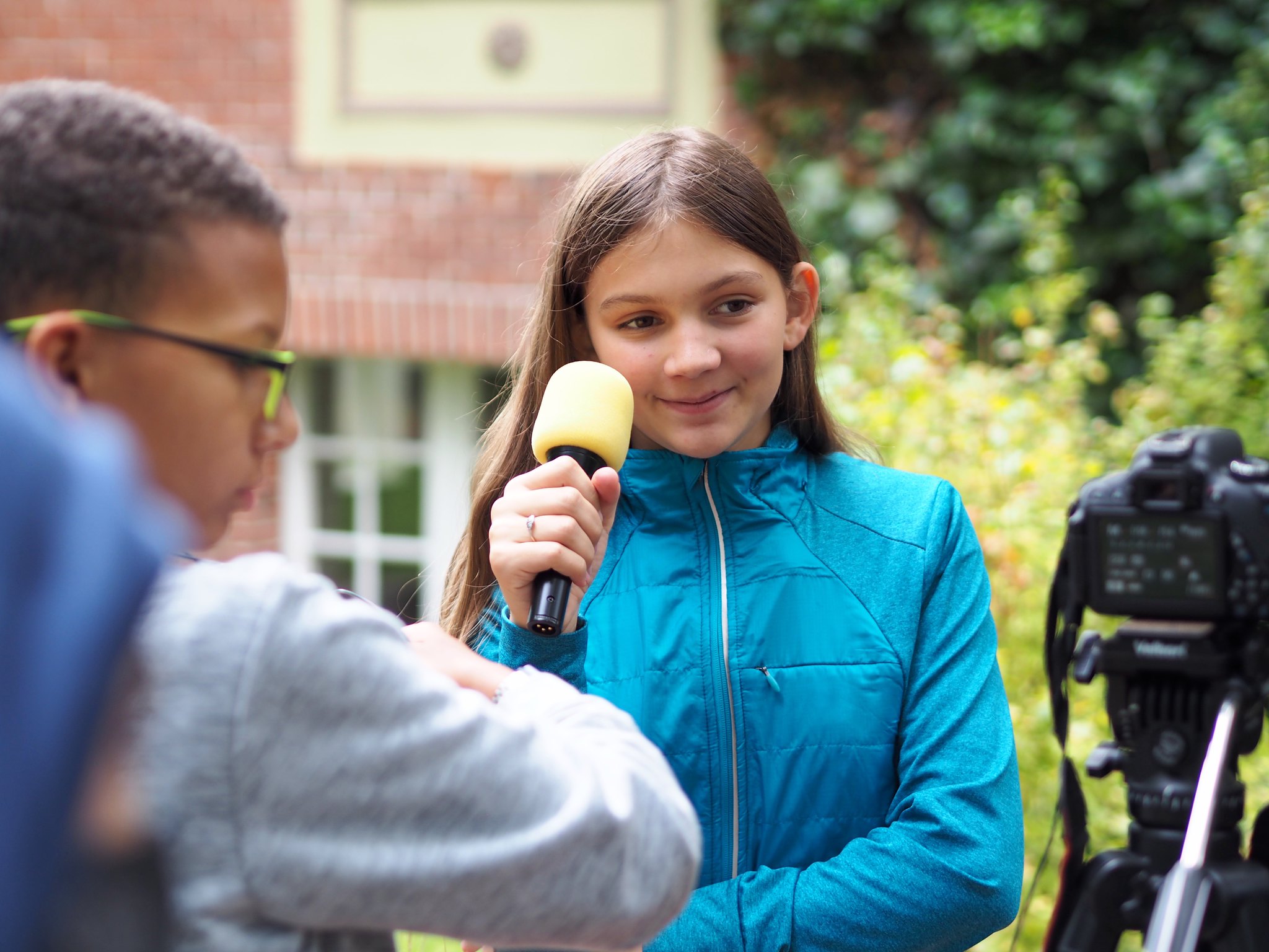 Mädchen mit blauer Jacke hält Mikrofon vor Kamera bei Interview im Freien