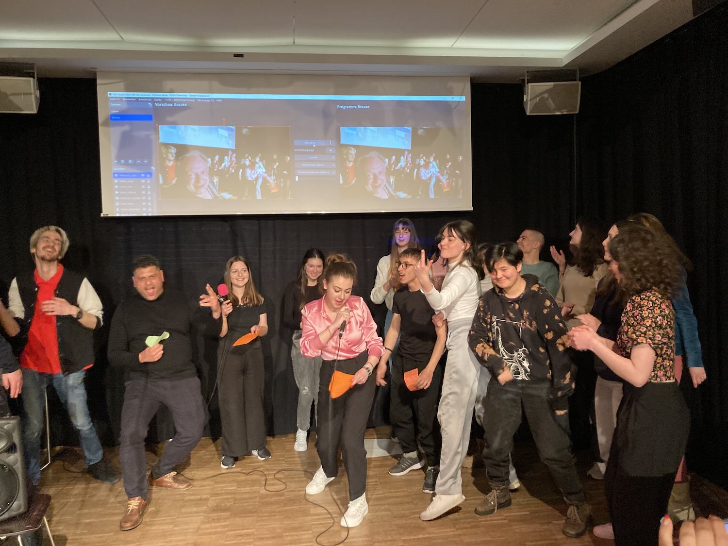 Gruppe von Jugendlichen auf Bühne bei internationaler Scheibwerkstatt 2023, einige halten bunte Zettel, im Hintergrund Projektion mit Fotos