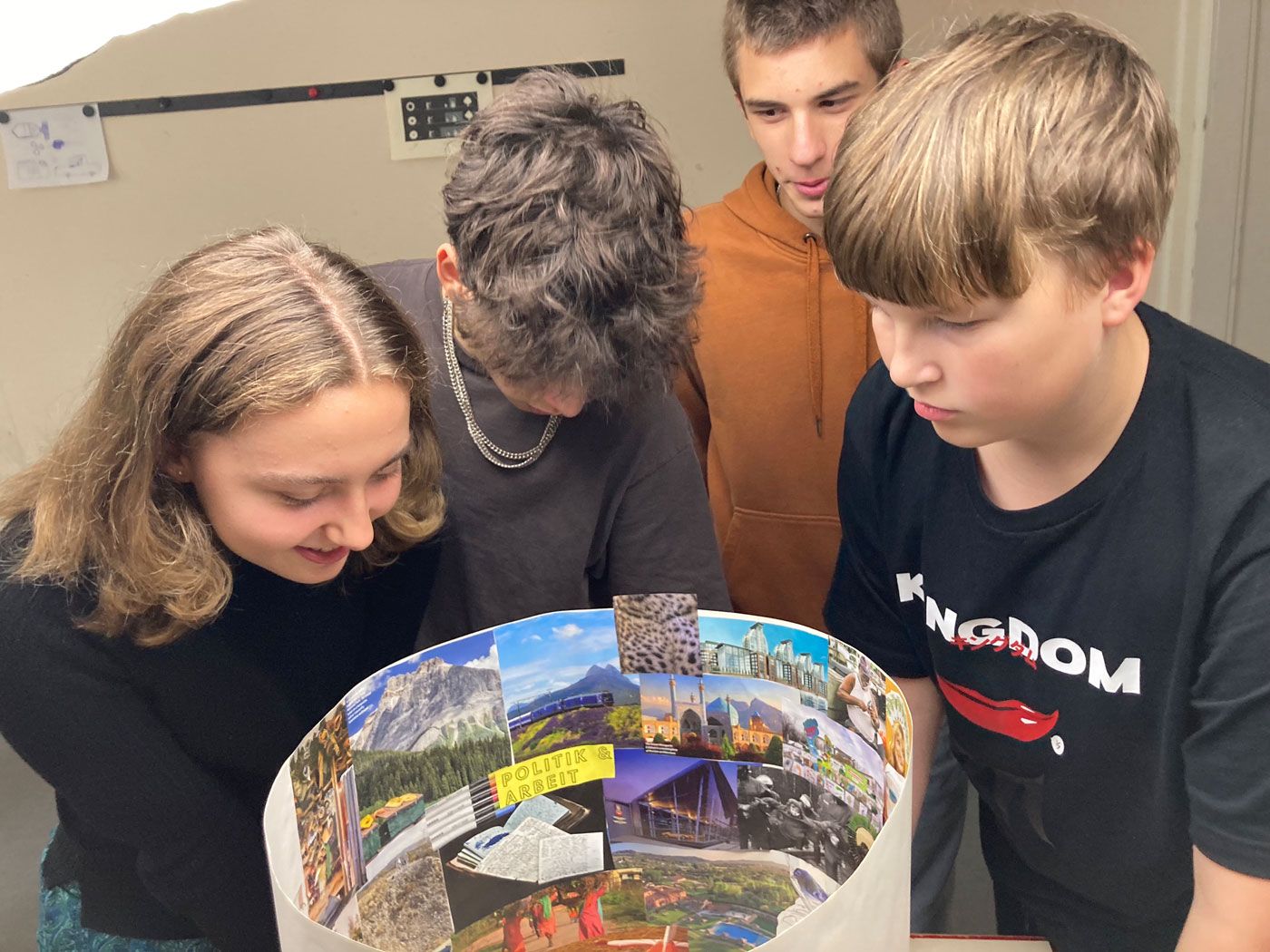 Vier Jugendliche blicken auf ein rundes Plakat mit Fotos von Landschaften, Gebäuden und Texten wie 'POLITIK'