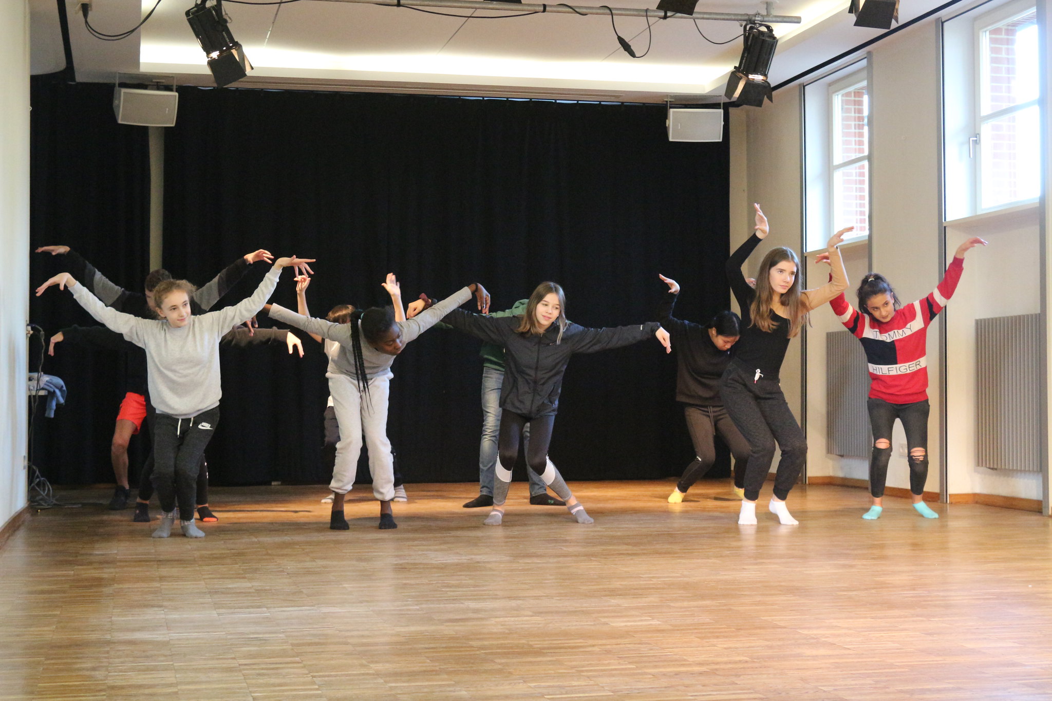 Gruppe von Jugendlichen tanzt synchron mit ausgebreiteten Armen in einem Tanzstudio mit Holzfußboden und schwarzem Vorhang