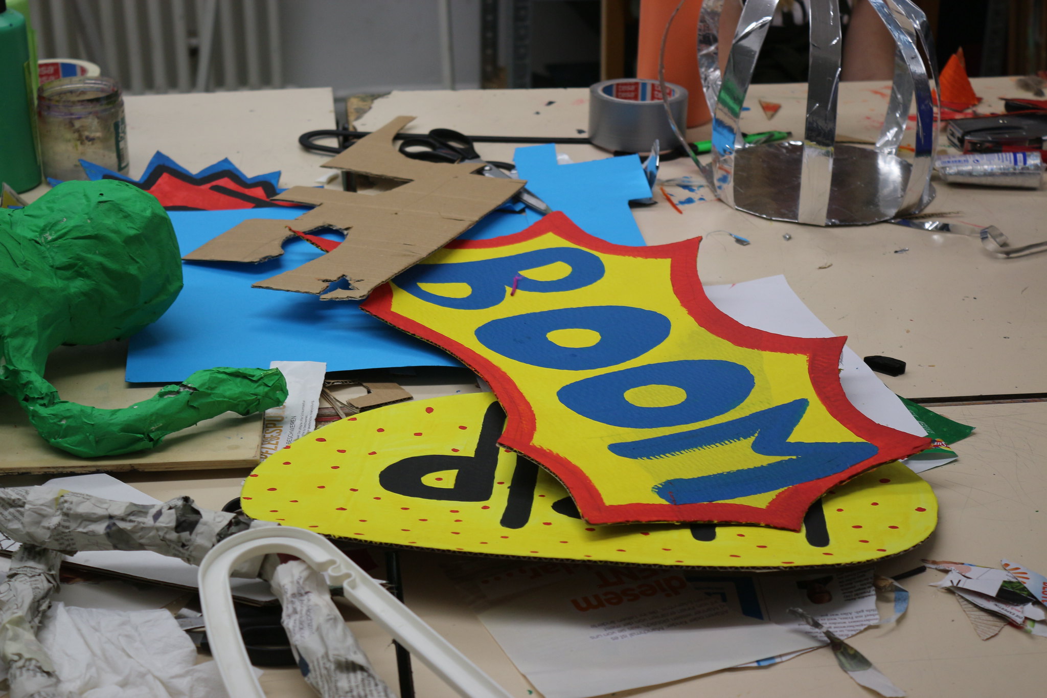 Bunte Pappschilder mit Comic-Texten 'BOOM' und 'POP' auf einem Tisch mit Bastelmaterialien