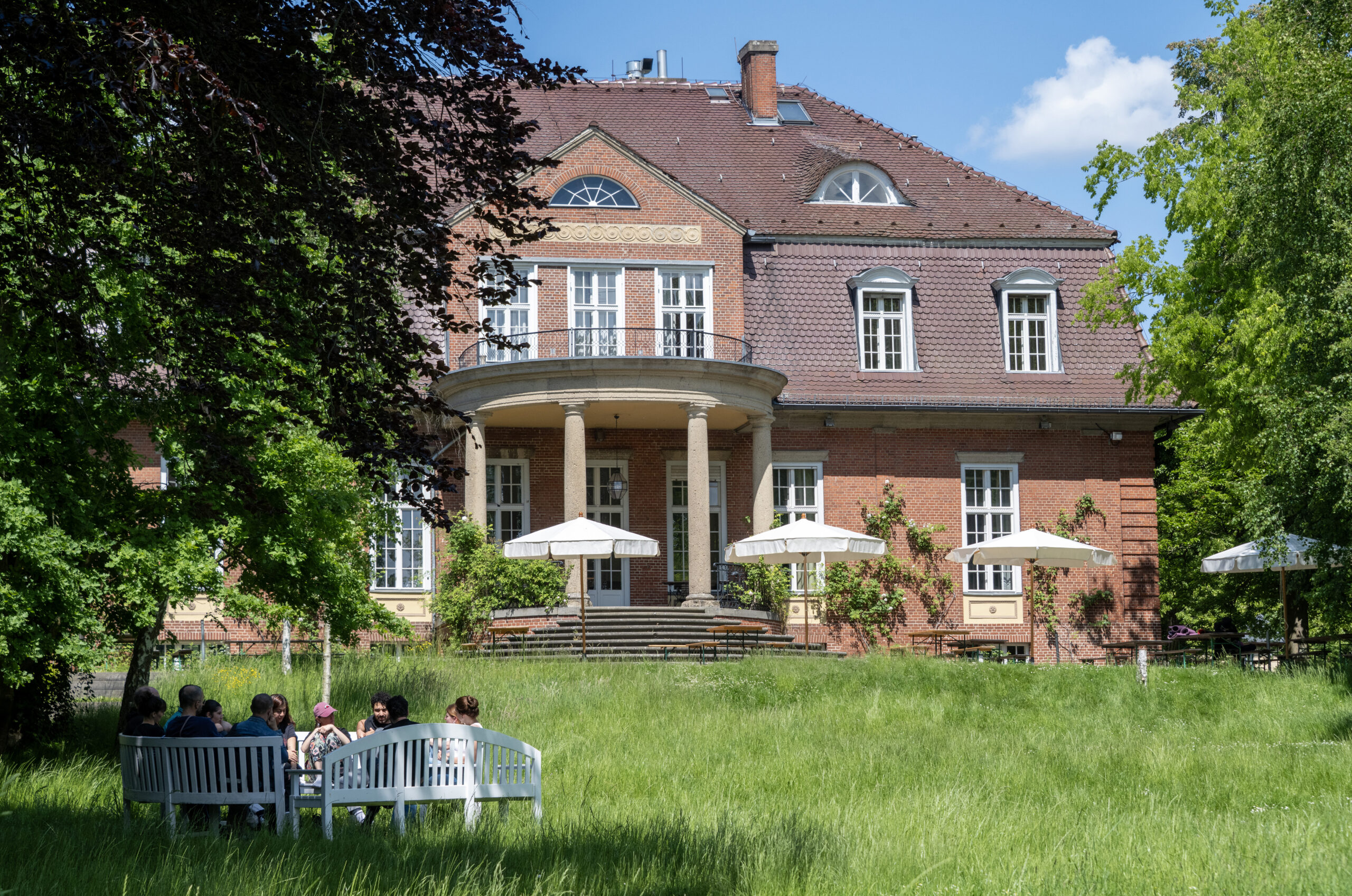 A group of people are sitting on white benches in the garden in front of a large red brick house with columns and two parasols