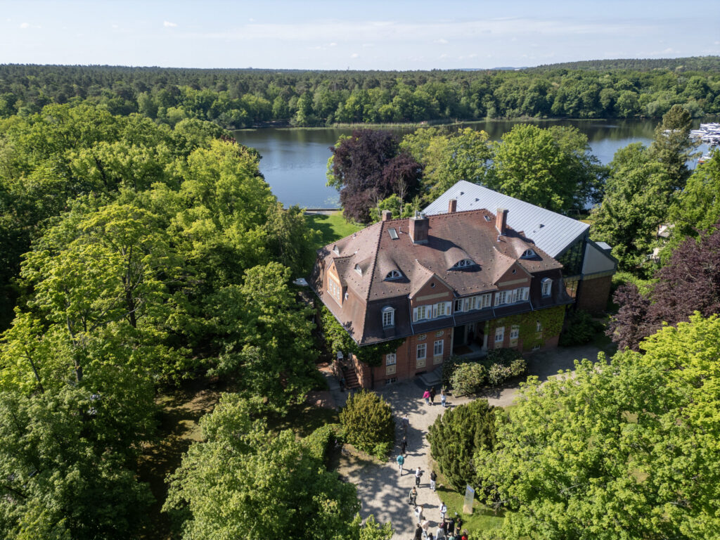 Historic brick Landhaus with several dormer windows, surrounded by trees, on the shore of a lake with forest in the background