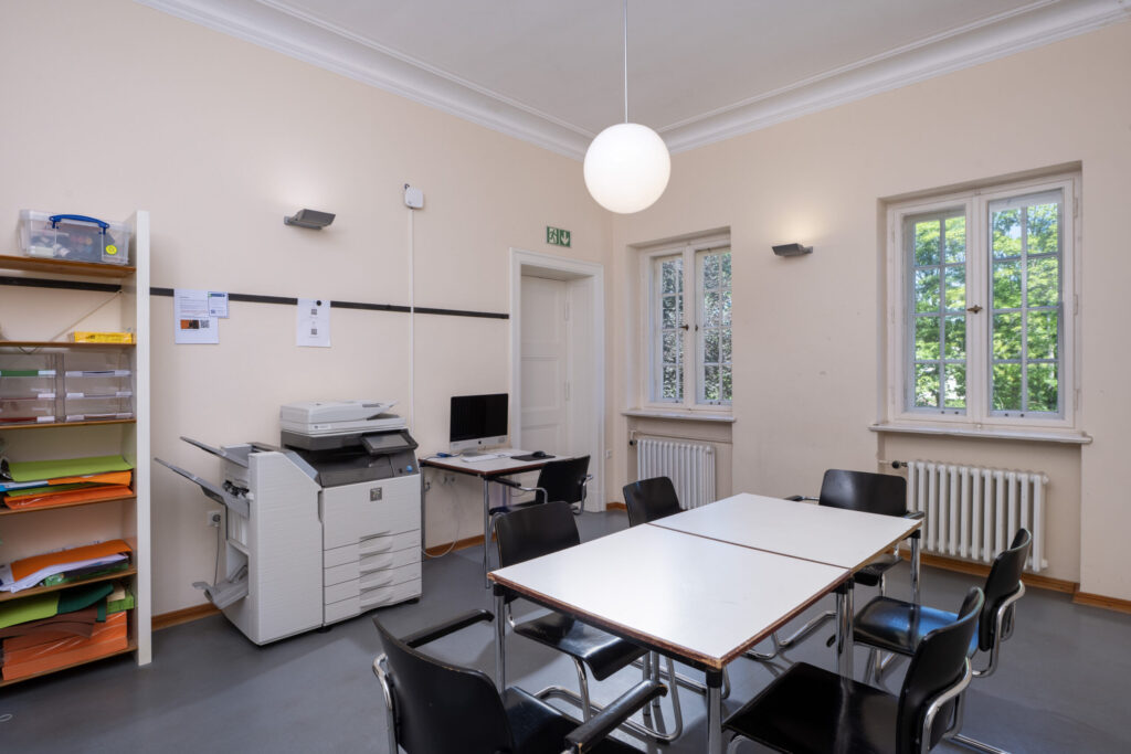 Seminarraum Elbenzimmer mit sechs schwarzen Stühlen um zwei zusammengestellte weiße Tische, Drucker und Computer an der Wand, Fenster mit Blick ins Grüne.