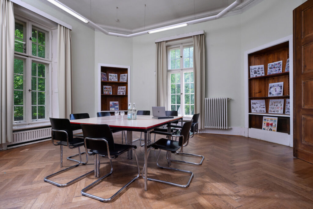 Seminarraum Gartenzimmer mit großem Fenster, quadratischem Tisch und sechs schwarzen Stühlen auf Parkettboden.