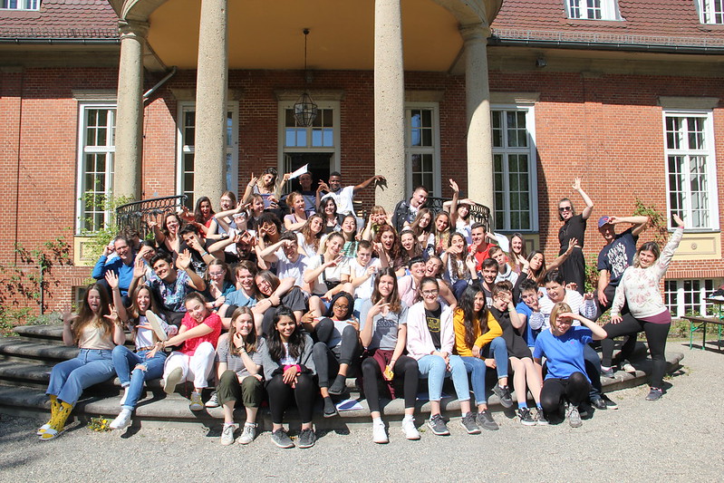Large group of young people sitting and standing on steps in front of a brick building with columns, many waving or making gestures
