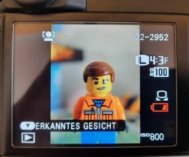 The digital camera displays a close-up of a Lego figure wearing orange clothing and a blue tie, marked as a recognized face