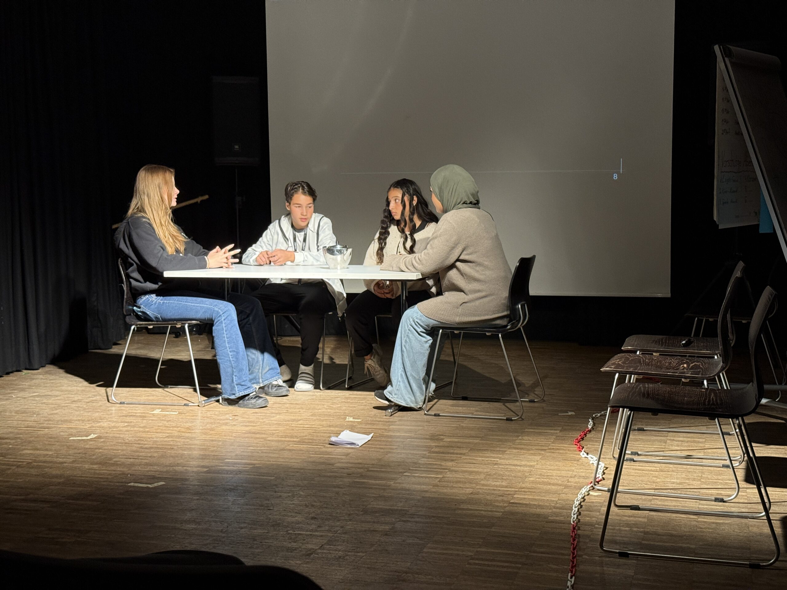 Four people are sitting at a white table on chairs in a dark room with a wooden floor and a canvas in the background.