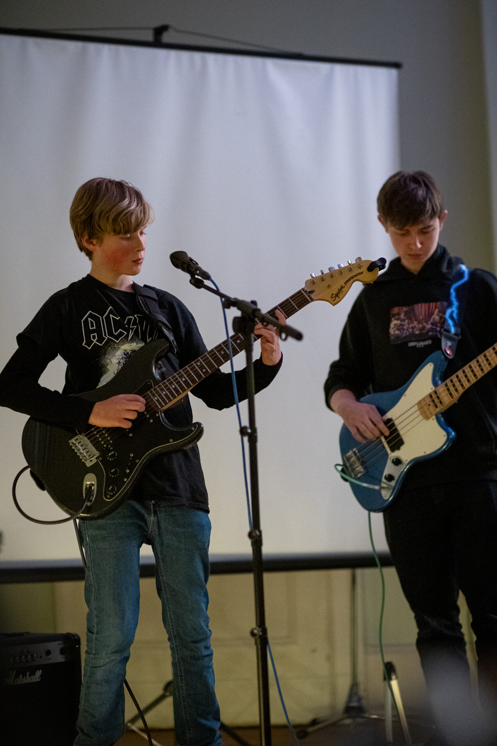 Two teenagers are playing electric guitar and electric bass in front of a screen, both wearing dark tops and jeans.