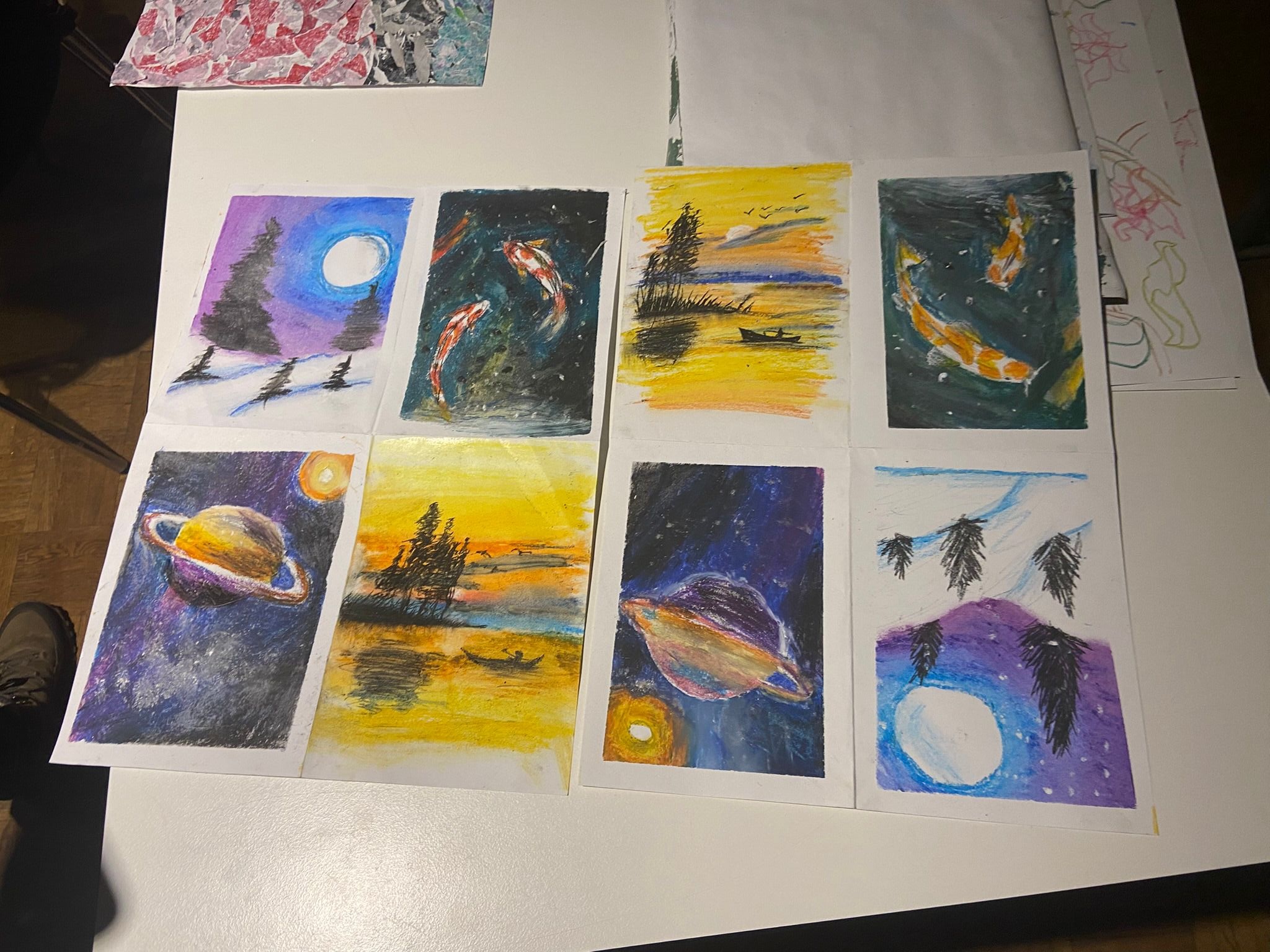 Acht bunte Aquarell- oder Pastellbilder auf einem weißen Tisch, darunter Darstellungen von Planeten, Fischen, Landschaften mit Bäumen und einem Boot sowie einem Mond mit Zweigen im Vordergrund.