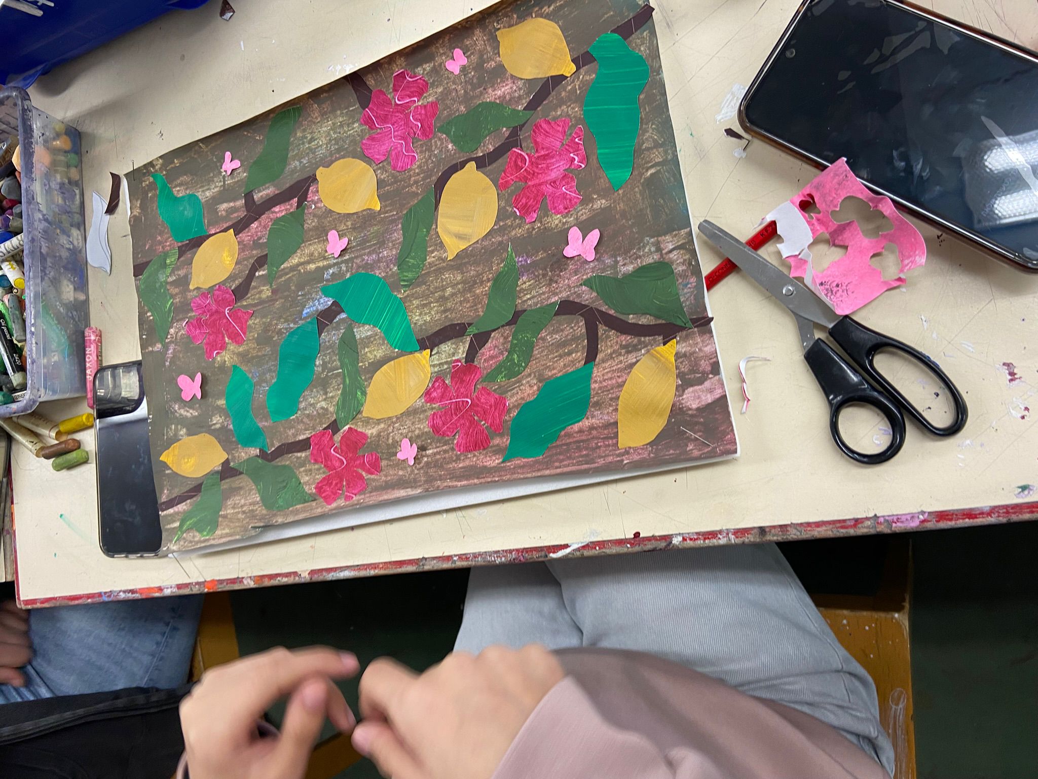 Arbeit mit buntem Papier, das grüne Blätter, gelbe Blätter und pinke Blumen zeigt, auf einem Tisch mit Schere, Smartphone und Händen im Vordergrund