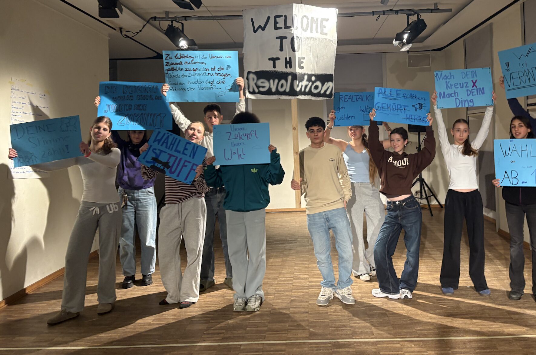 Eine Gruppe Jugendlicher steht in einem Raum. Jede Person trägt ein Protestschild.