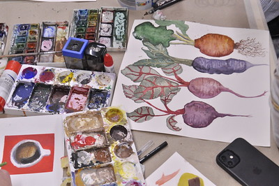 Auf dem Tisch liegen Dinge zum Malen. Farbkästen, Pinsel. Auf einem Bild sieht man verschiedene Wurzelgemüse.