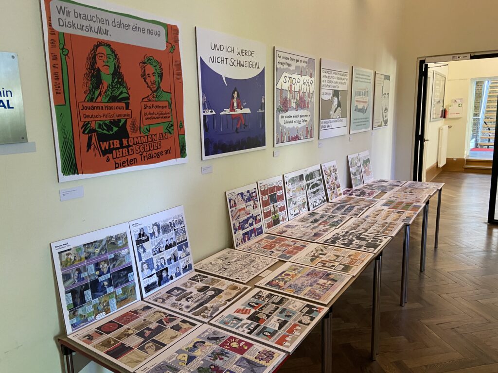 Ausstellung mit mehreren Comic- und Plakatwerken an einer Wand und auf Tischen in einem hellen Raum mit offenem Durchgang