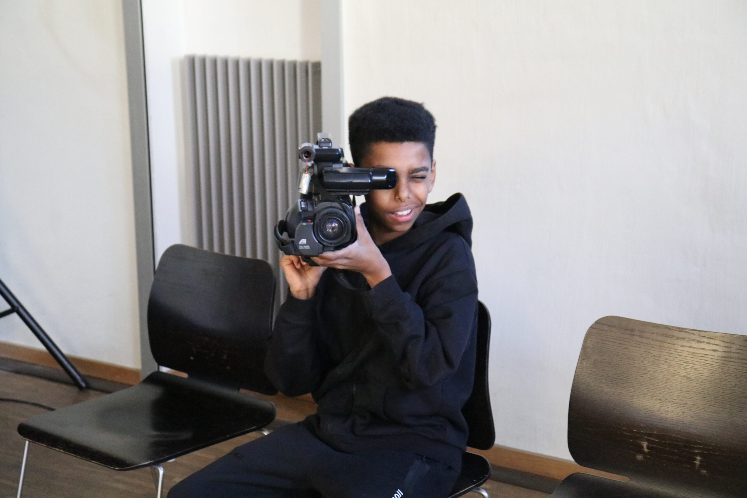 Ein Junge im schwarzen Jogginganzug sitzt und hält eine Videokamera vor sein Gesicht.