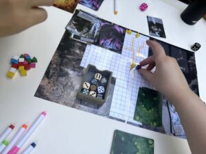 Zwei Hände bewegen Spielfiguren und Würfel auf einem Spielplan mit Karten, Würfeln, bunten Markern und Stiften.