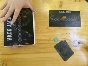 Eine Hand mit langen, lackierten Fingernägeln hält ein aufgeschlagenes Spielheft neben einem Spielbrett mit der Aufschrift 'HACK JACK', Spielfigur und Karten auf einem Holztisch.