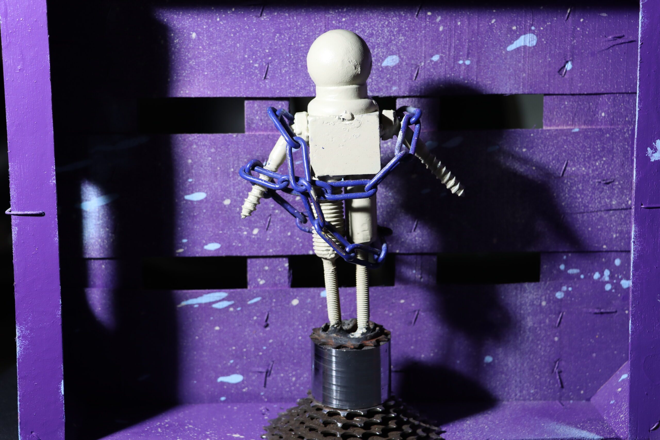 Eine Weiße Metall Figur in einer lilanen Box die in eienr Kette liegt. Die Figur wurde aus unterschiedlichen Teilen wie Schrauben zusammen geschweißt.