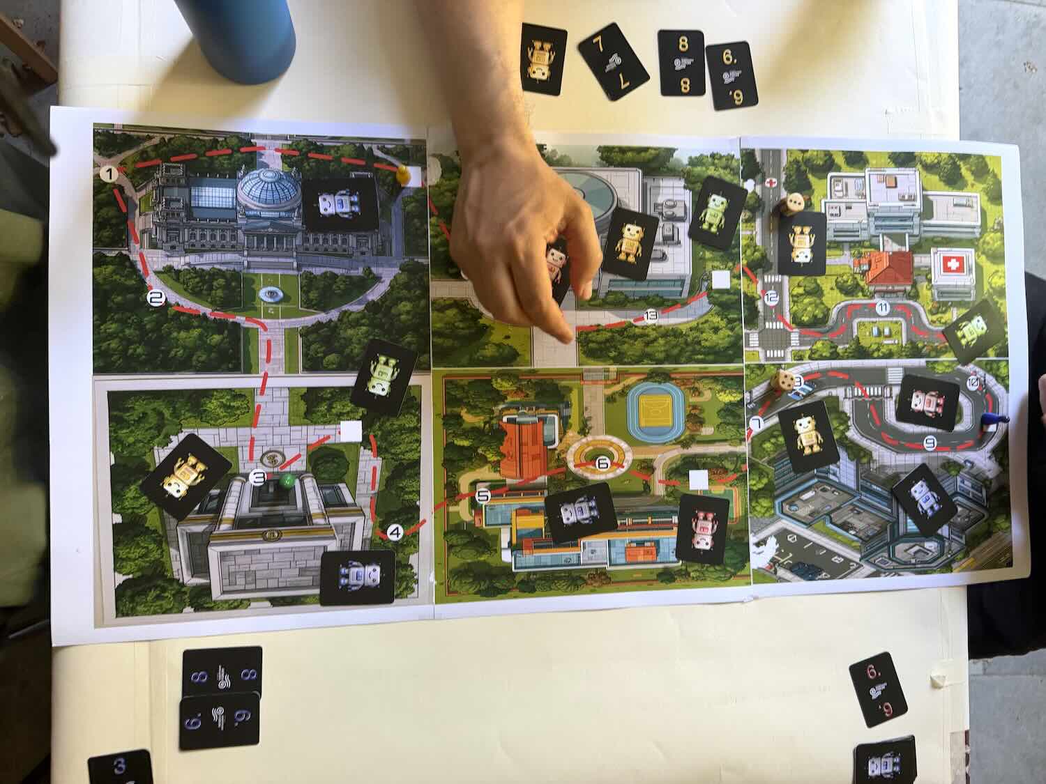 Eine Hand legt eine Spielkarte auf einen Spielplan mit mehreren Gebäuden und weiteren ausliegenden Karten.
