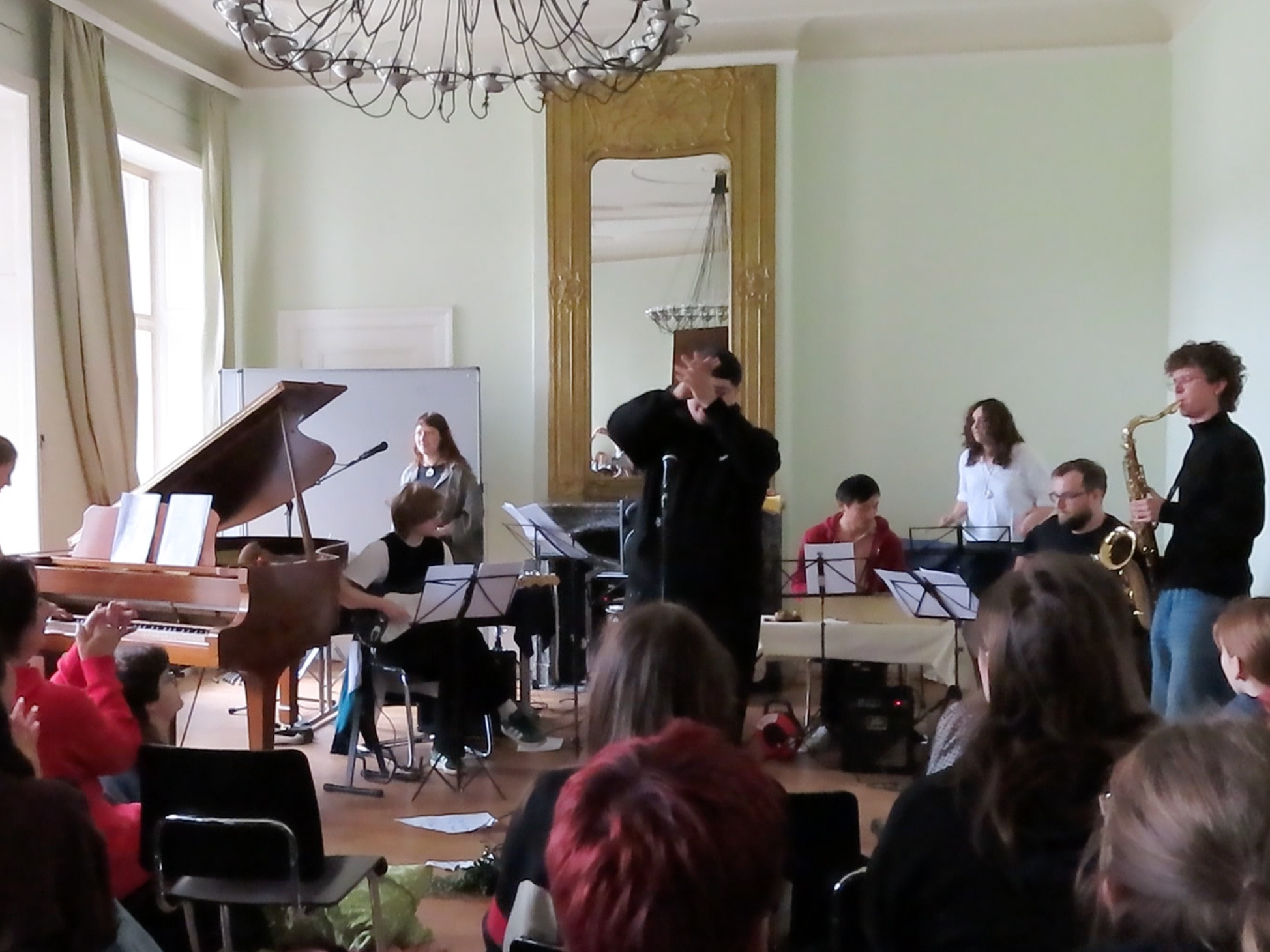 Mehrere Musiker spielen in einem Raum mit Flügel, Saxofon und weiteren Instrumenten vor einem Publikum