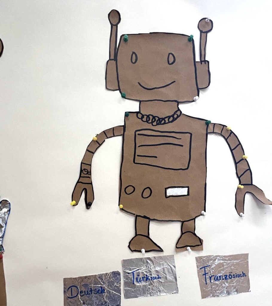 Aus Pappe ausgeschnittener Roboter mit rechteckigem Kopf, zwei Antennen, lächelndem Gesicht, rechteckigem Körper mit Linien und Kreisen, sowie einfachen Armen und Beinen, an einer Wand befestigt