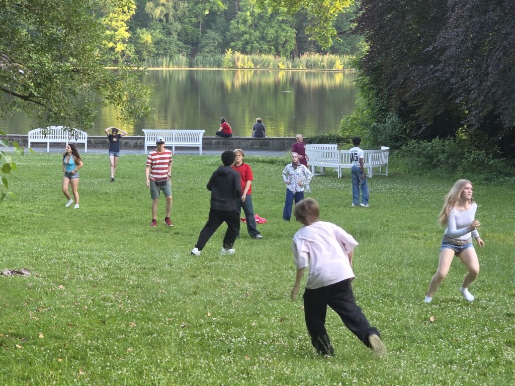 junge Menschen spielen und rennen auf einer Wiese am See.