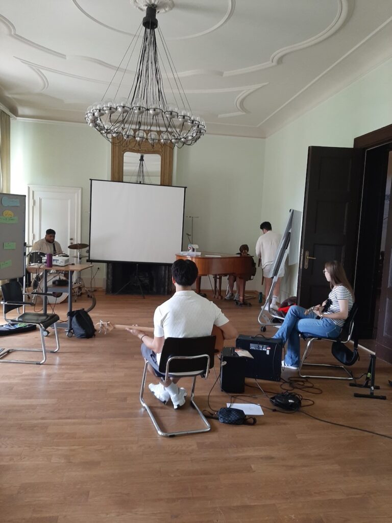 Mehrere Personen sitzen mit Musikinstrumenten und Verstärkern in einem Raum mit einem Klavier, einer Leinwand und einem Kronleuchter.