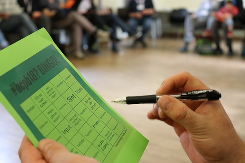 Hand hält einen grünen Zettel mit einer Bingo-Tabelle und einem Stift, im Hintergrund sitzen mehrere Personen unscharf auf Stühlen.