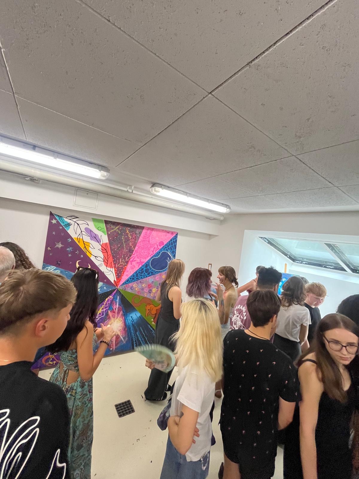 Gruppe junger Menschen steht in einem Raum vor einer bunten, geometrisch gemusterten Wandmalerei mit verschiedenen Farben und Formen