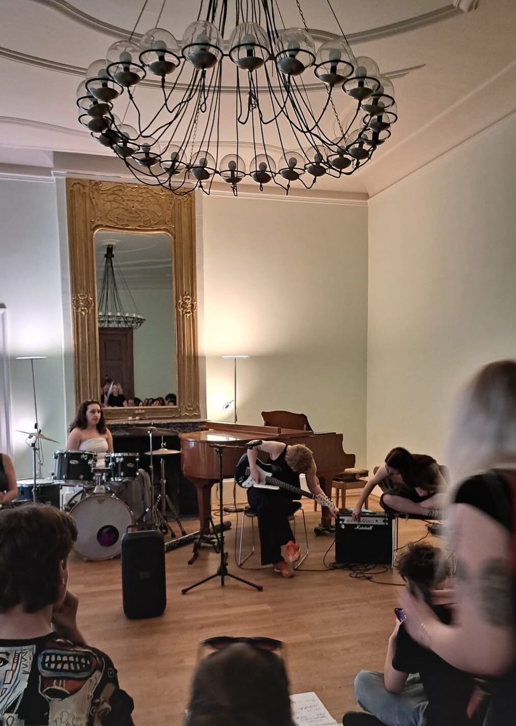 Musiker spielen in einem Raum mit großem Spiegel und Kronleuchter vor sitzendem Publikum