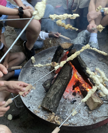 Mehrere Personen halten mit Stöcken umwickelte Teigstücke über ein offenes Lagerfeuer