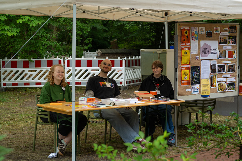 Drei Personen sitzen an einem Tisch unter einem Pavillon vor einer Stellwand mit Flyern und Plakaten im Freien