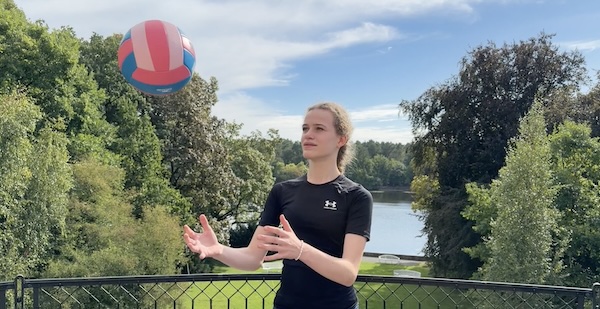 Junge Frau in schwarzem Sportshirt fängt einen rot-blau-weißen Ball auf einer Terrasse mit Blick auf einen See und Bäume