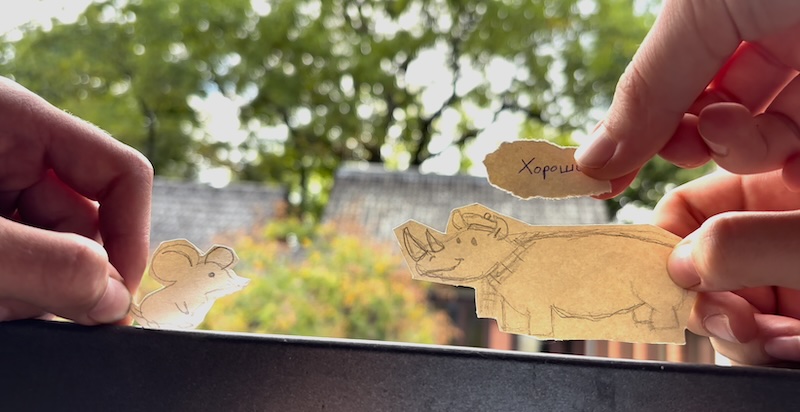 Zwei Hände halten ausgeschnittene Papiertiere, eine Maus links und ein Nashorn rechts, vor einem unscharfen Hintergrund mit Bäumen und Häusern
